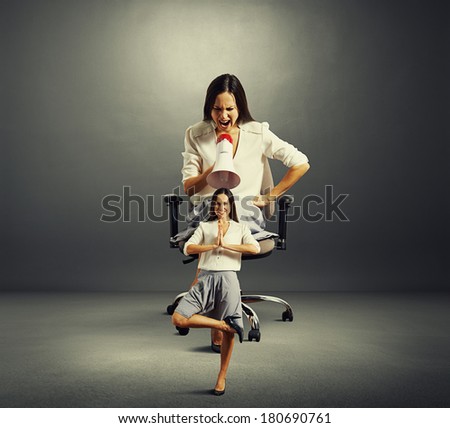 angry woman sitting on the office chair and screaming at small calm woman over dark background