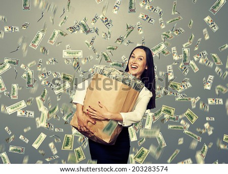 excited young businesswoman standing under dollar\'s rain and holding big paper bag with money