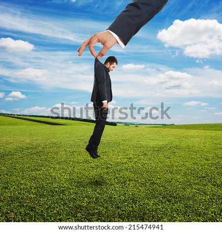 big hand holding small sad businessman in suit over green field and blue sky