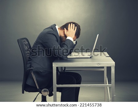 depressed businessman sitting at the table with laptop and covering his head over dark background