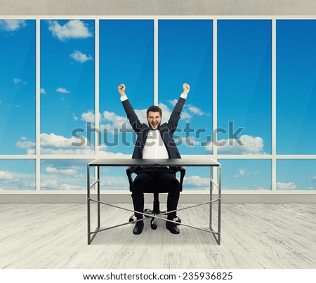 happy businessman sitting at the table, raising hands up up and laughing in the light office with big windows