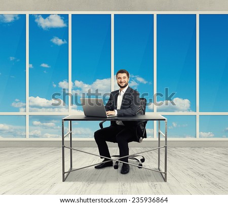 smiley young businessman sitting at the table with laptop and looking at camera in the light office with big windows