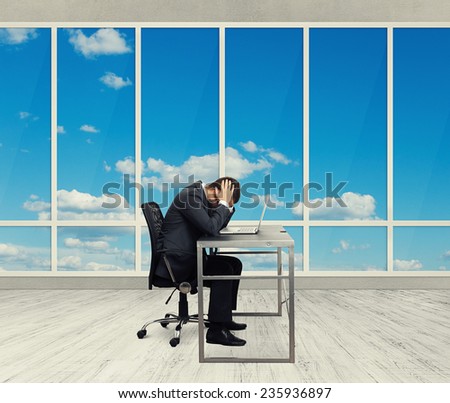 depressed businessman sitting at the table with laptop and covering his head in the light office with big windows