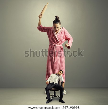 angry big wife with rolling pin screaming at lazy small husband in the grey room