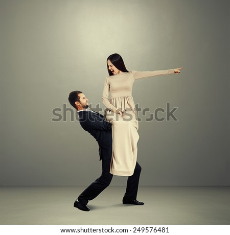 dissatisfied young woman in long dress sitting on tired man, pointing at something and screaming. photo over grey background