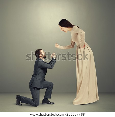 full length portrait of emotional couple over grey background. woman screaming and showing fist, man standing on knee and apologizing