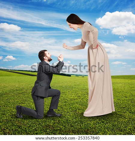 full length portrait of emotional couple at outdoor. woman screaming and showing fist, man standing on knee and apologizing