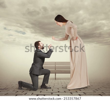 full length portrait of emotional couple in foggy park. woman screaming and showing fist, man standing on knee and apologizing
