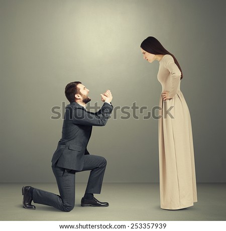 full length portrait of emotional couple over grey background. angry woman looking at man, man standing on knee and apologizing