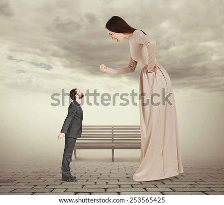 angry screaming woman showing fist and looking down at small kissing man. concept photo in foggy park