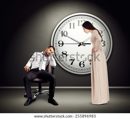 angry woman staring at lazy man on chair. photo in empty dark room with big white clock on the wall
