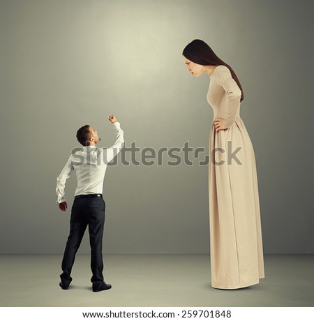 small angry man showing fist to dissatisfied woman in long dress over grey background
