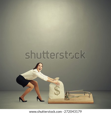 excited woman in formal wear reaching her arms to take bag with money in big mousetrap over dark grey background with empty copyspace overhead