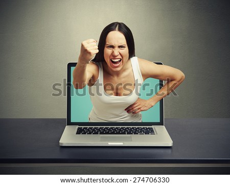 angry screaming woman stretching out of laptop and showing fist at camera against dark background