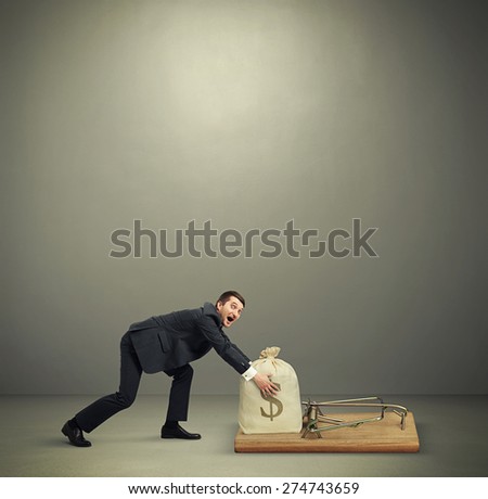 amazed man in formal wear reaching his hands to take bag with money in big mousetrap over dark grey background with empty copyspace overhead