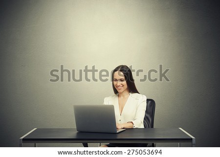 smiley woman sitting at the table with laptop, typing on keyboard and looking at screen over dark grey background with empty copyspace overhead