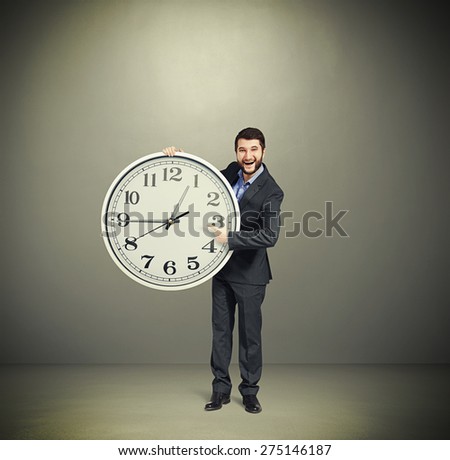 laughing successful businessman in formal wear pointing at clock dial and looking at camera over dark grey background