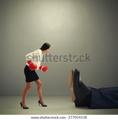 angry businesswoman in formal wear and boxer gloves looking at big knocked out man over dark grey background