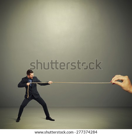 businessman in formal wear and big womans hand pulling the rope over dark grey background