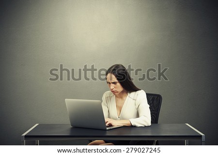 serious woman sitting at the table with laptop, typing on keyboard and looking at screen over dark grey background with empty copyspace overhead