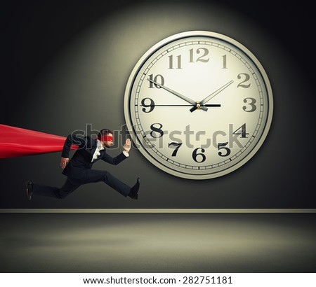 serious businessman wearing like superman running against big white clock in dark room