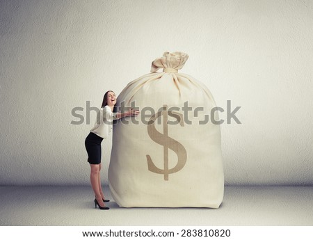 happy woman embracing big bag with money and smiling in empty grey room