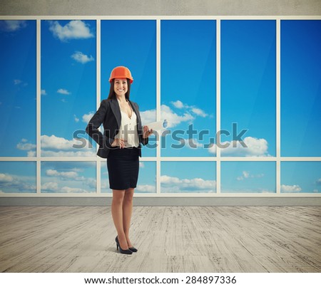 smiley woman architect in orange hardhat holding plan and looking at camera in room with big windows