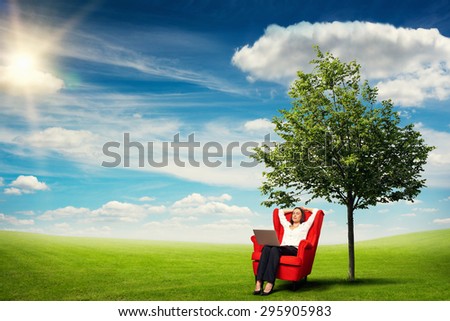 smiley businesswoman with laptop on knees relaxing on the red chair over beautiful landscape with green meadow, tree and blue sky