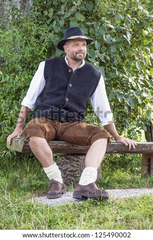 Seated man in traditional Bavarian costumes on a wooden bench in front of bushes