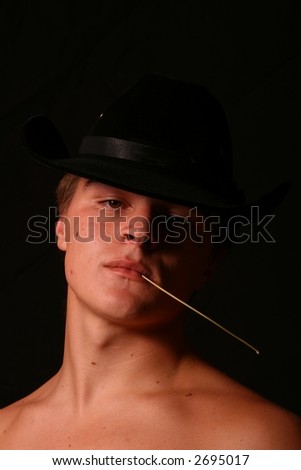 Shirtless male model in Cowboy Hat chewing on a grass stem casting a shadow over his face looking at the camera