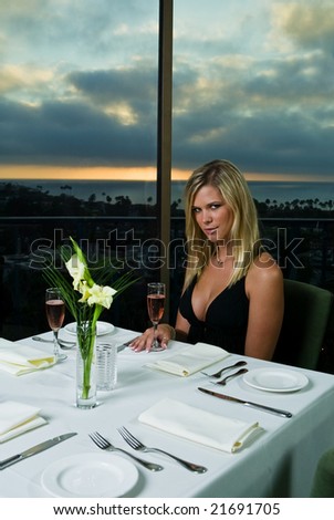A beautiful blond woman in a seductive black dress having dinner at a nice restaurant with the sun setting in the background