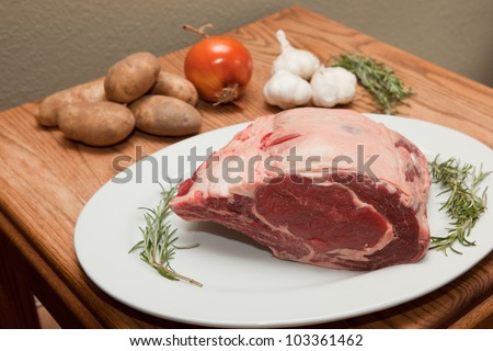 Seven pound bone-in prime rib beef roast, uncooked on a white plate garnished with onion, garlic, rosemary and potatoes.