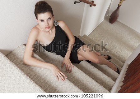 Beautiful, elegant feminine woman sitting on the steps indoors, wearing a black evening gown, showing some leg