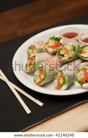 Diagonal view of shrimp spring rolls with sweet chili sauce and chopsticks on placemat with two-toned wood tabletop.
