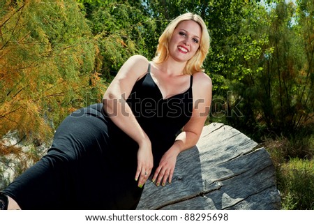 Beautiful full figured blonde woman wearing black dress resting on a log. She is wearing cowboy boots, and it is autumn.