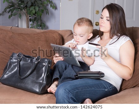 Young mother shopping online with her credit card on her tablet while her toddler son sits on her lap.