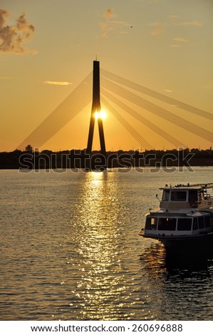 Tourist boat floats on the river at sunset.\
The Shroud Bridge (Latvian: VanÅ¡u tilts) in Riga is a cable-stayed bridge that crosses the Daugava river in Riga, the capital of Latvia.