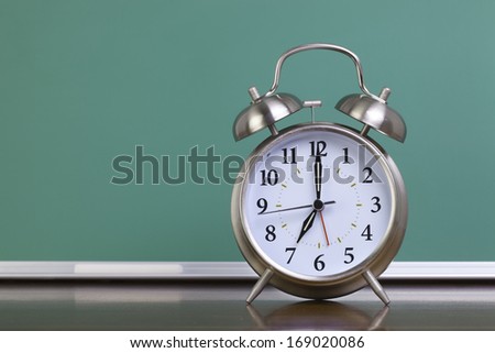 A silver alarm clock in front of a green chalkboard