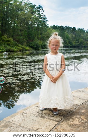 Five-year girl in a beautiful white dress standing near a pond overgrown with duckweed