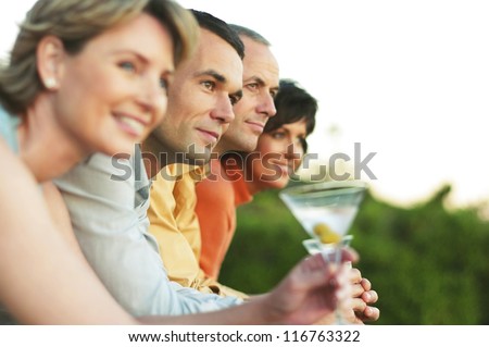 Four middle-aged friends stand in a row leaning on a rail enjoying martini cocktails