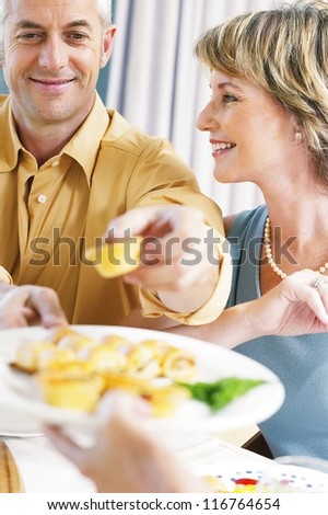 Attractive natural couple at the dining table with the husband stretching forwards to help himself to appetizers, focus to faces.