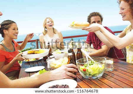 A happy group of teenage friends sitting at a table at the seaside drinking beer and eating their lunch