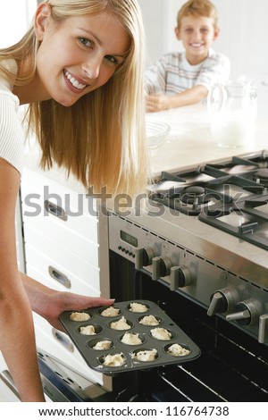 Smiling young housewife baking a tray of cookies which she is placing in the oven watched in the background by her son