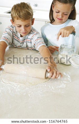 Little boy l rolling pastry in the kitchen with a rolling pin as his sister sprinkles fresh flour to prevent it sticking