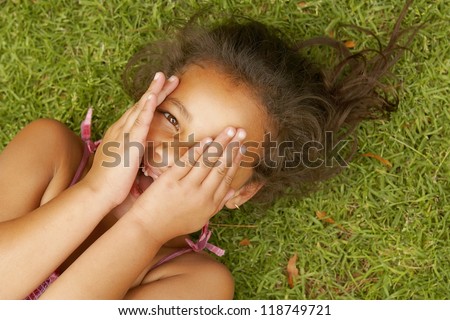 Little girl lying on the green grass on her back playing peek a boo with the camera peering out between her fingers which are covering her face