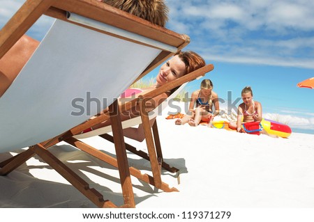 Young couple relax in deck chairs while their small daughters play nearby in the white sand on a summer beach