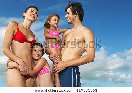 Young family with two small daughters posing close together in the swimsuits against a blue sky on a happy summer holiday