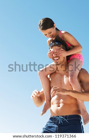 Little girl covers her dads eyes as she is carried on his shoulders while playing at the beach