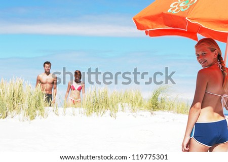 Young girl with large orange beach umbrella smiling back over her shoulder at the camera as her parents approach across the sand dunes