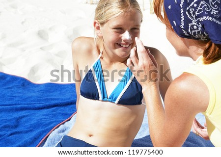 Mother applying suntan lotion to the nose and face of her young daughter who is stretched out on a towel in her bikini at the beach